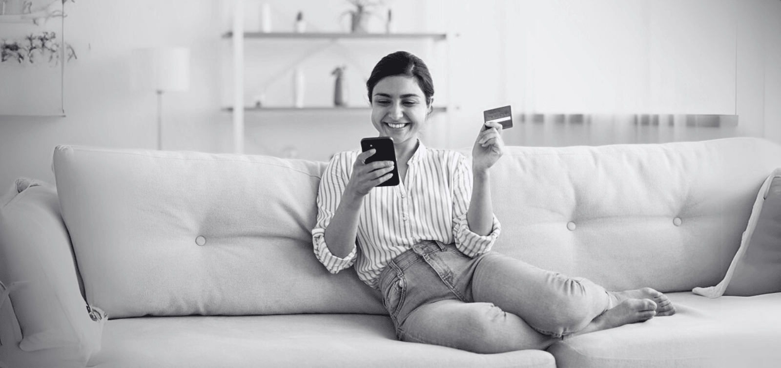 Young adult female holding a Credit Builder credit card and looking at her phone while at home on the couch.
