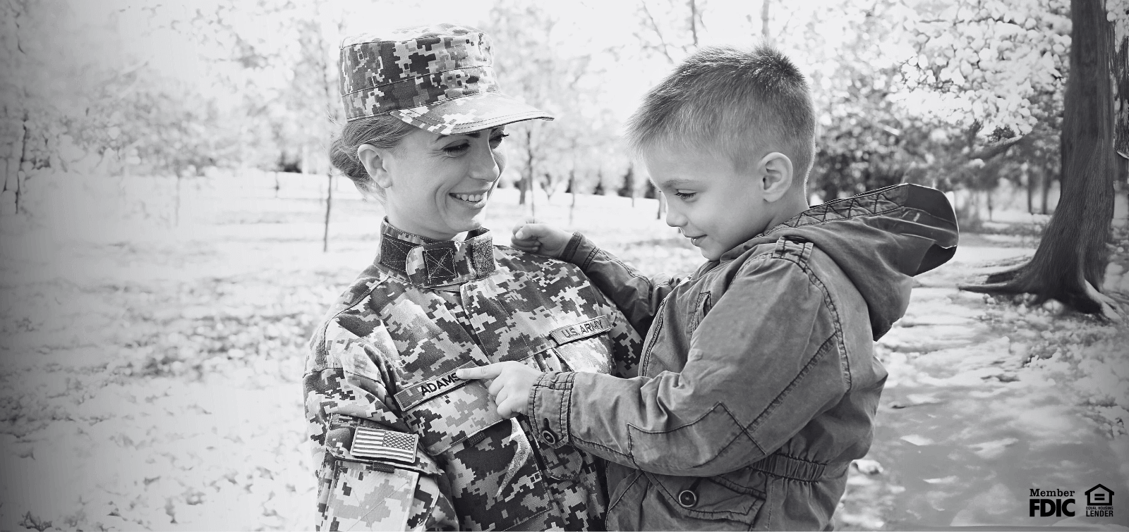 A female soldier smiles while holding a young child on a sunny day. 